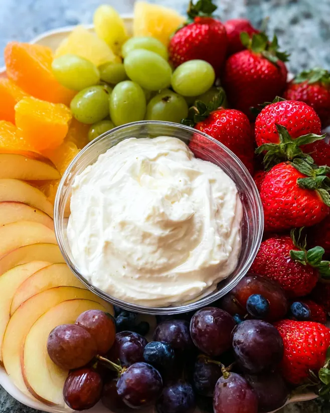 A bowl of cream cheese dip sits on a table with a variety of fruits.