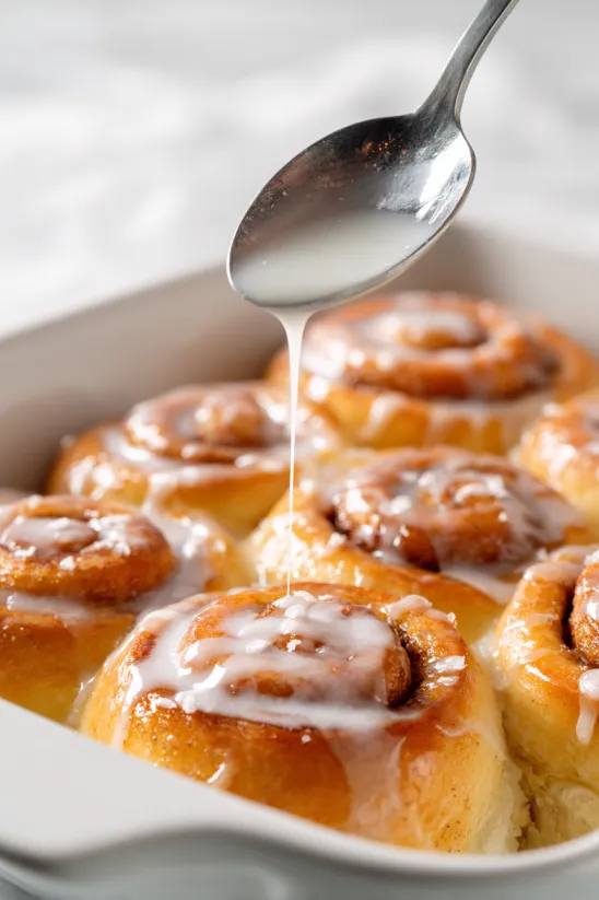 A spoon is drizzling icing over a plate of cinnamon rolls.