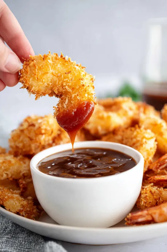 A person is dipping a piece of coconut shrimp into a bowl of sauce.