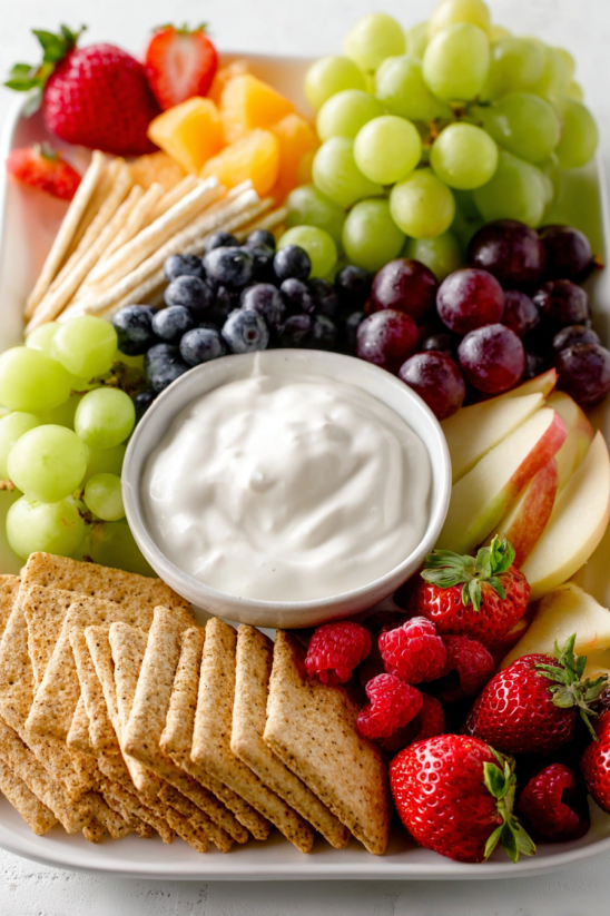 A bowl of cream cheese sits on a plate with fruit.