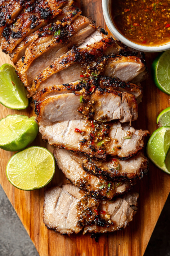 A plate of lechon asado with limes and a bowl of sauce.