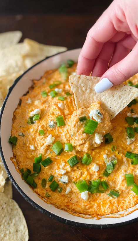 A hand is grabbing a tortilla chip from a bowl of slow cooker buffalo chicken dip.