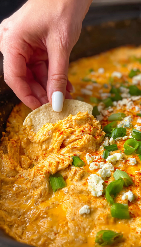 A hand is dipping a tortilla chip into a slow cooker buffalo chicken dip.